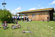 Ein einfaches einstöckiges Holzhaus mit Satteldach, das eine Dachbegrünung hat. Davor Kinder-Fahrräder die im Rasen abgestellt wurden und eine Gruppe Menschen, die sich unter dem Vordach versammelt hat. Ganz links ein Maibaum und Sträucher.
