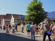 Menchen gehen auf einer Straße bergauf entlang von Fachwerkhäusern, welche zum Teil Photovoltaik auf dem Dach haben. Hinter einer Mauer steht ein großer Laubbbaum.