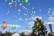 Viele bunte Luftballons werden auf einem Dorfplatz von Bürgern in den Himmel aufsteigen gelassen.