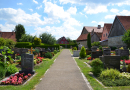 Ein Weg führt durch einen Friedhof mit dunklen Grabsteinen und unterschiedlich bepflanzten Gräbern.