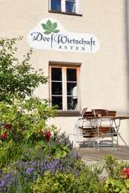 Ein nach traditioneller Art ocker-beige verputztes Bauernhaus, man sieht ein unbehandeltes Lärchenholzfenster und darüber einen handgemalten Schriftzug „Dorfwirtschaft“. Direkt vor dem Fenster grenzt eine Holzterrasse an, auf der eine Gartentischgarnitur steht. Im Vordergrund eine schöne Pflanzung mit Rosen und Lavendel.